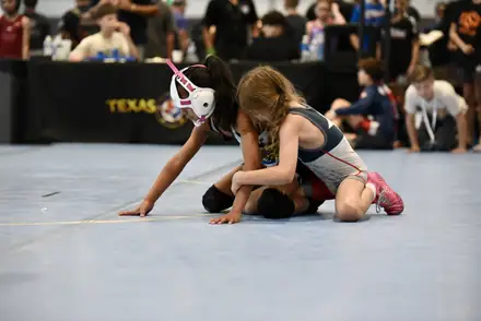 A young girl in headgear controls her opponent on the mat at a state tournament