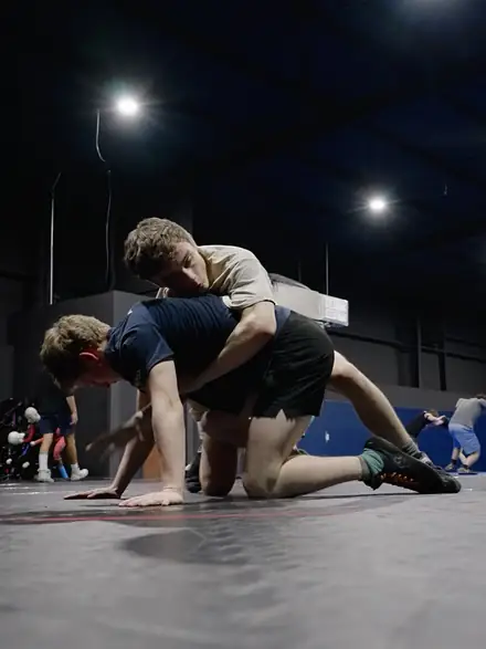 Two wrestlers scrambling for position on the mat under the gym lights.