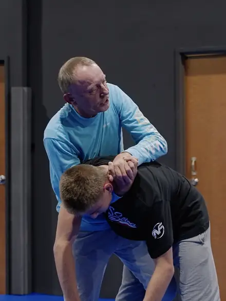 A coach in a light blue shirt walking a young wrestler through a collar tie.