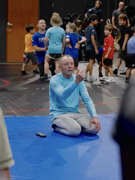 A coach kneeling on the mat to teach while a group of young wrestlers listens.