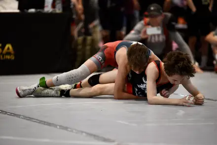 An AWC wrestler in orange and black rides an opponent at a USA Wrestling tournament