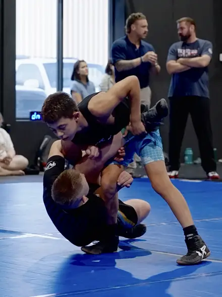 A wrestler lifting a partner through a drill while coaches watch from the side.